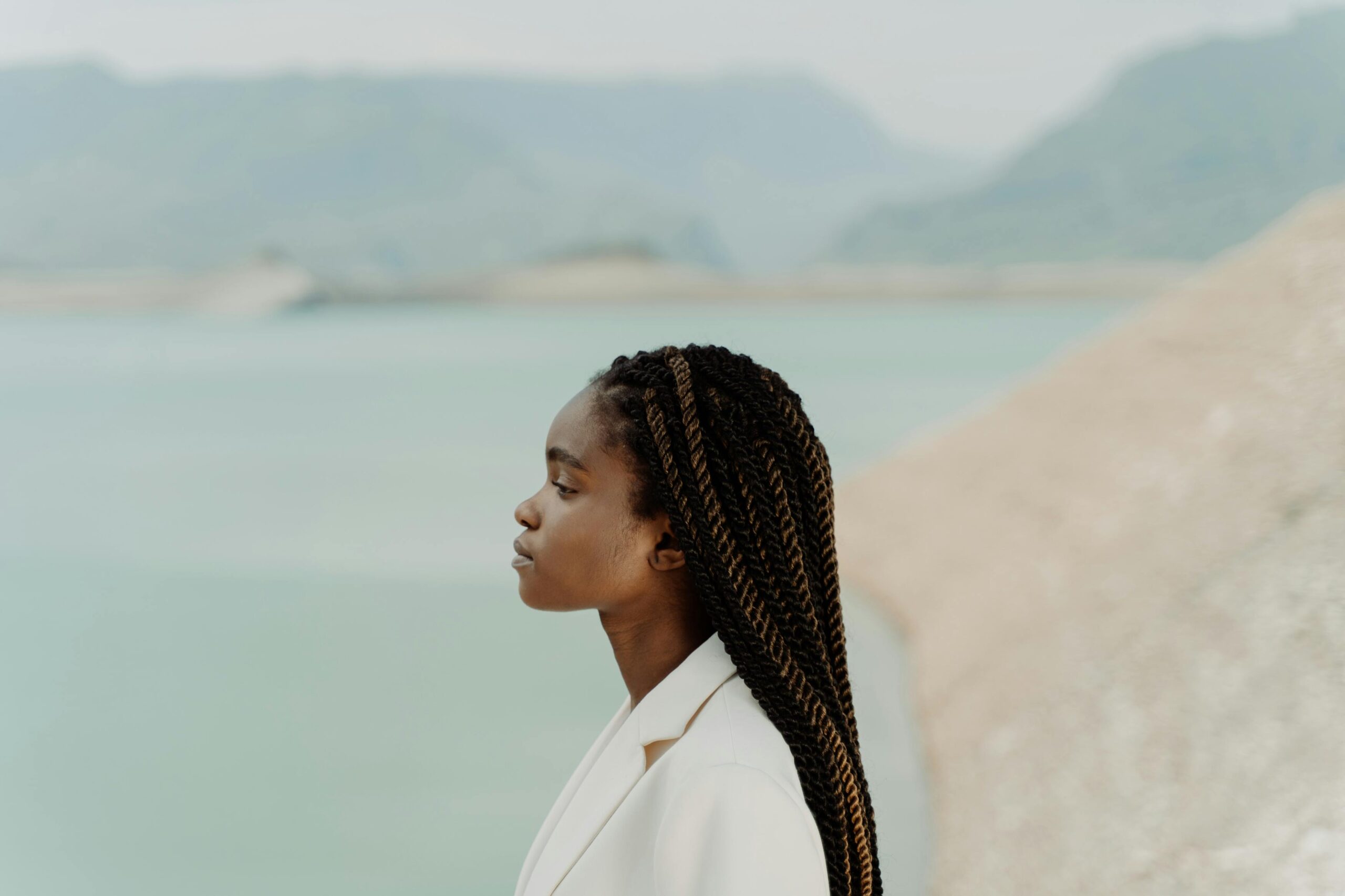 Profile portrait of a stylish woman with afro braids in a white suit by a serene lake.